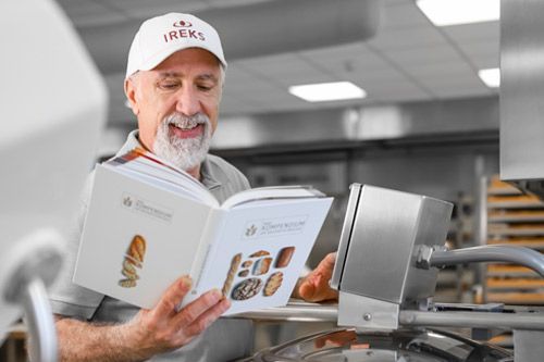 Ein Bäcker steht in der Bäckerei und liest im IREKS-Kompendium der Bäckereitechnologie